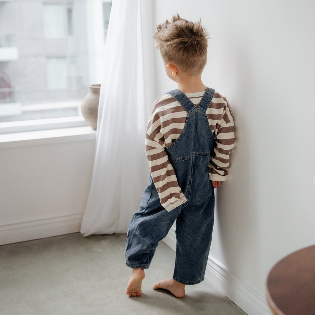 Child wearing a striped shirt and denim overalls standing near a window.