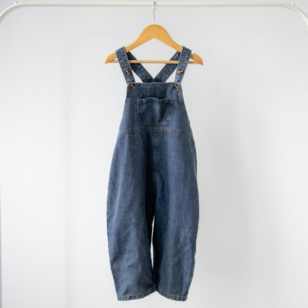 Blue denim overalls hanging on a wooden hanger against a white background