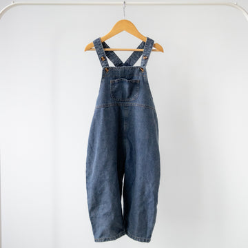 Blue denim overalls hanging on a wooden hanger against a white background