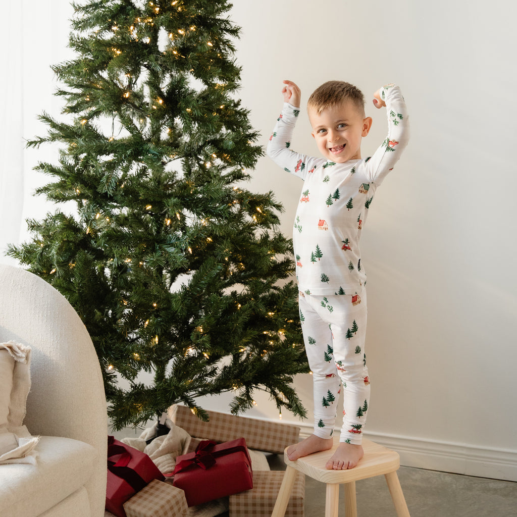 Child in pajamas standing on a stool next to a decorated Christmas tree with presents underneath.